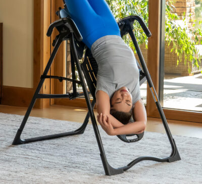 A woman is inverted on a teeter inversion table, demonstrating a stretching exercise for back relief.