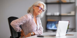 Older woman sitting at a desk, slouched posture, highlighting the impact of bad posture on back pain.