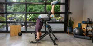 Person performing an inversion exercise on a Teeter DEX II fitness machine in a modern home gym, surrounded by greenery and fitness equipment, emphasizing core strength and flexibility training.