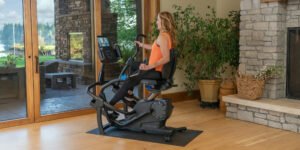 Woman exercising on a Teeter FreeStep recumbent elliptical machine in a bright, home fitness space with large windows and indoor plants.