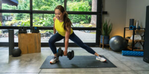 Woman performing a ProFlex dumbbell workout in a home gym, showcasing a lunge position on a workout mat. The background features fitness equipment, including weights, a stability ball, and a foam roller, with large windows revealing a lush outdoor garden.