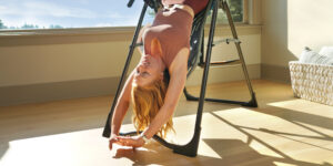 Woman using an inversion table for stretching and back relief in a sunlit room, promoting wellness and fitness.