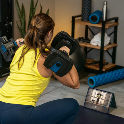 A person in workout clothes performs a squat with ProFlex adjustable dumbbells while following an exercise video displayed on a tablet in a home gym setting.