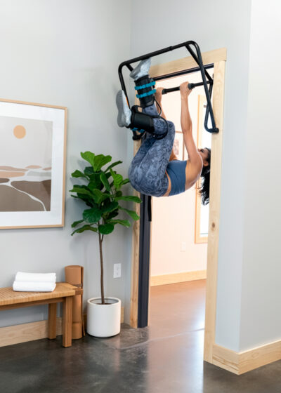 A woman hangs from a pull-up bar, using Teeter Gravity Boots, in a well-lit room with exercise equipment.