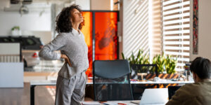 A woman stands in an office, stretching her back to relieve pain.